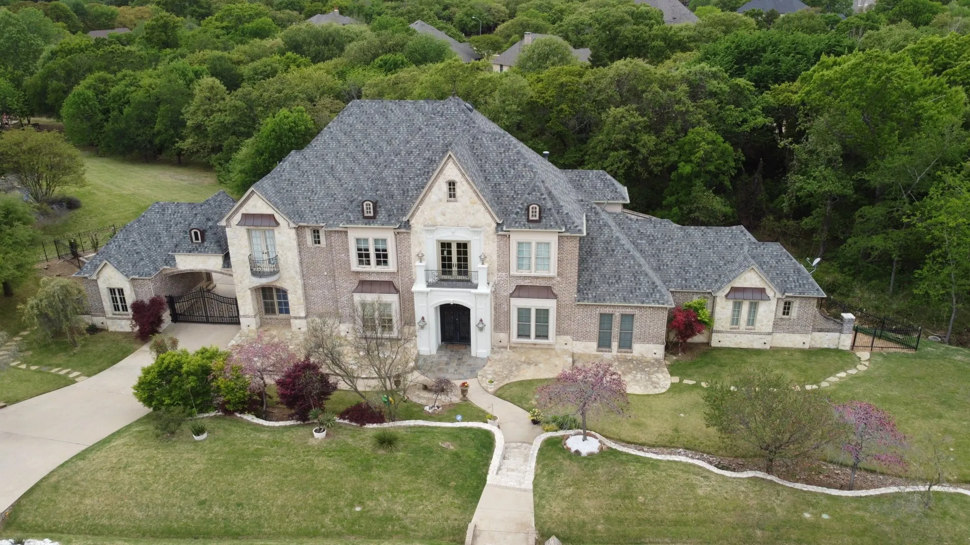 Large stone house with gray roof and landscaped front yard, surrounded by trees