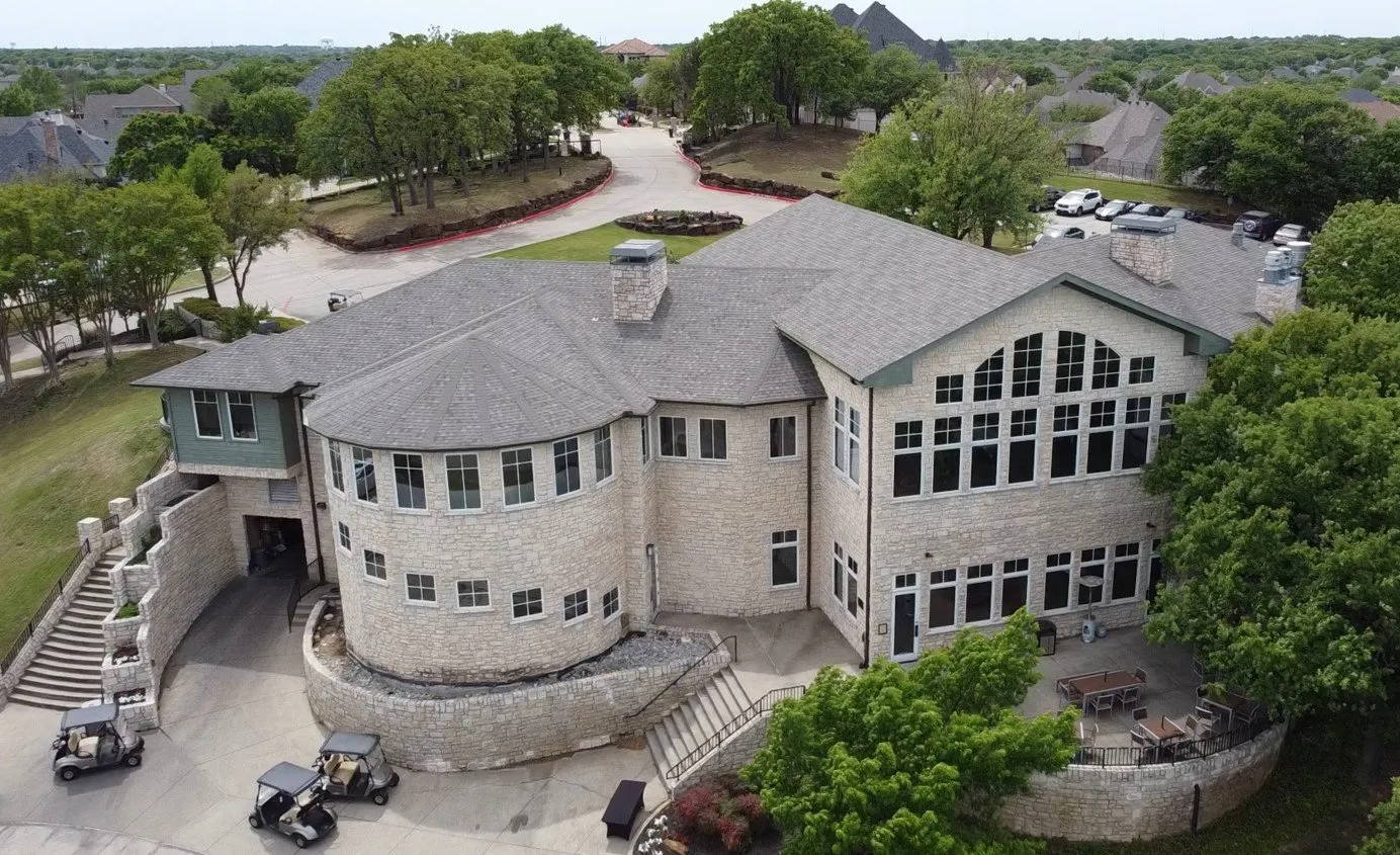 Aerial view of a large stone mansion with a gray roof, curved facade, and surrounding trees and driveway.